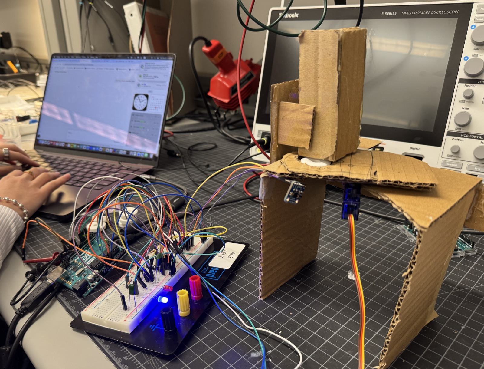 The full build on the bench: cardboard chassis, breadboard, oscilloscope, and Arduino, with the blue LED lit to show the system at rest.